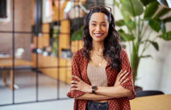 A person with long hair, wearing a red polka dot shirt and glasses on their head, stands smiling with arms crossed in a modern office with plants and a glass partition.