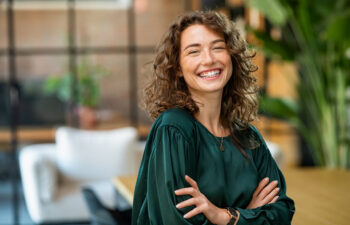 Woman with curly brown hair, wearing a green blouse, stands indoors with arms crossed and smiles at the camera. Modern office setting with plants and soft lighting in the background.