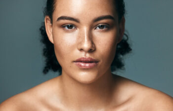 A young woman with curly hair and clear skin looks directly at the camera against a plain background, shoulders bare and expression neutral.