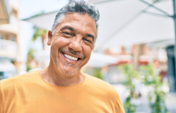 A smiling middle-aged man with gray hair and facial stubble wearing a bright yellow t-shirt, standing outdoors with blurred buildings and greenery in the background.