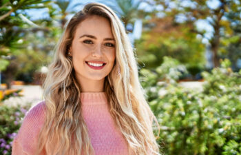 A woman with long blonde hair and a pink sweater smiles outdoors, with greenery and sunlight in the background.